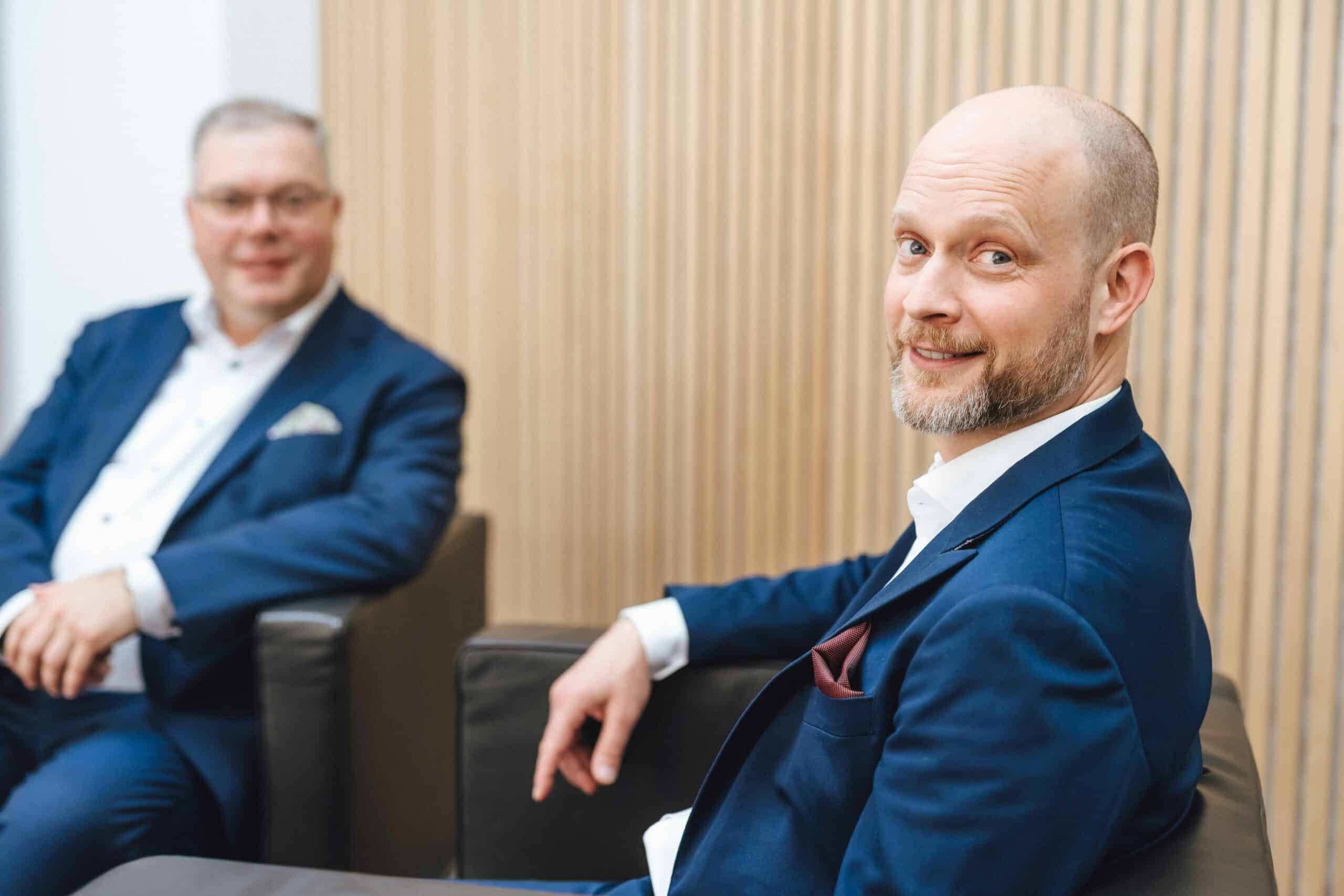 Dr. Carsten Walter, im blauen Anzug mit weißem Hemd, sitzt lächelnd in moderner Lounge, unscharfer Kollege im Hintergrund.
