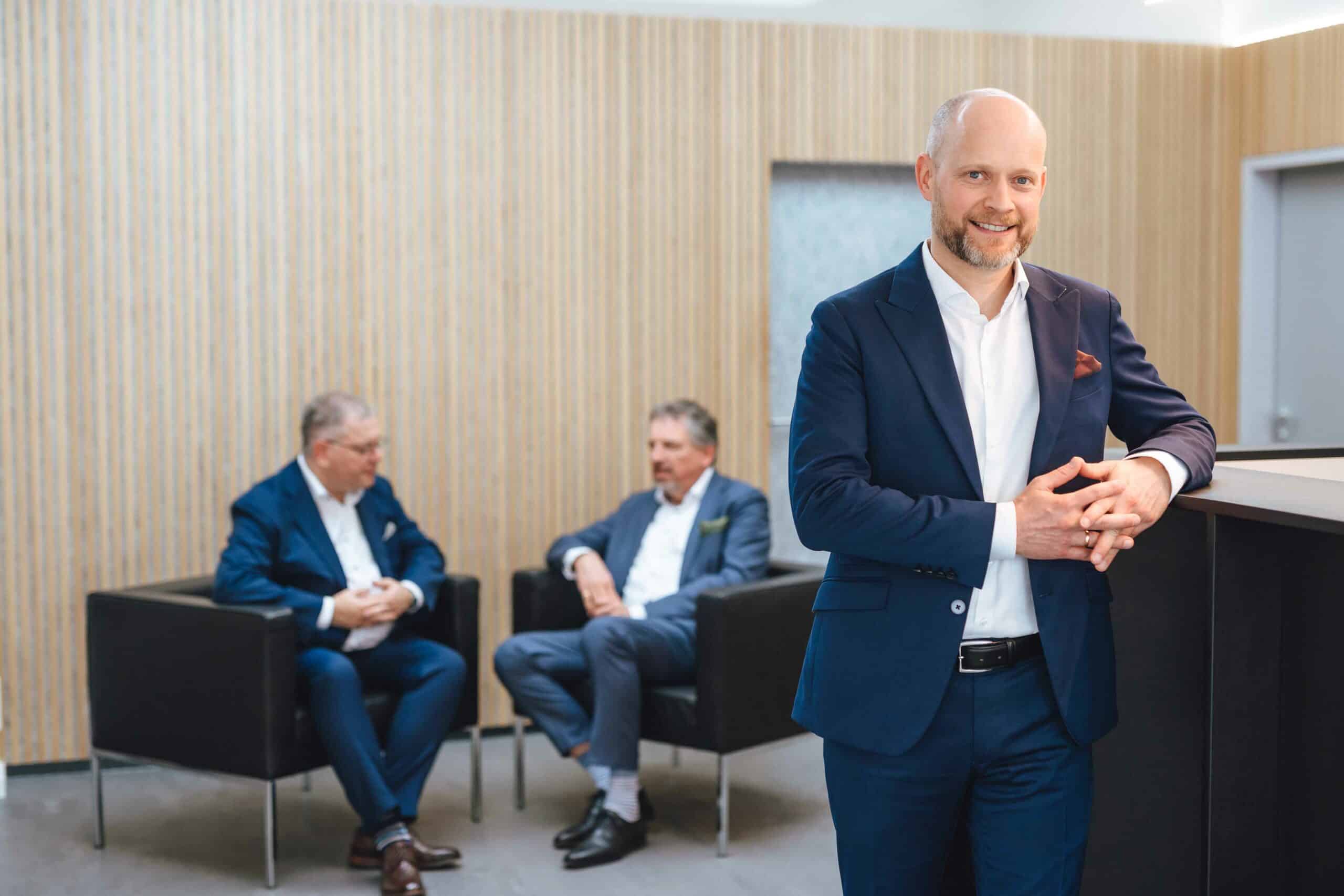 Dr. Carsten Walter, im blauen Anzug, lehnt an Tresen in moderner Kanzlei, zwei Kollegen im Hintergrund im Gespräch.