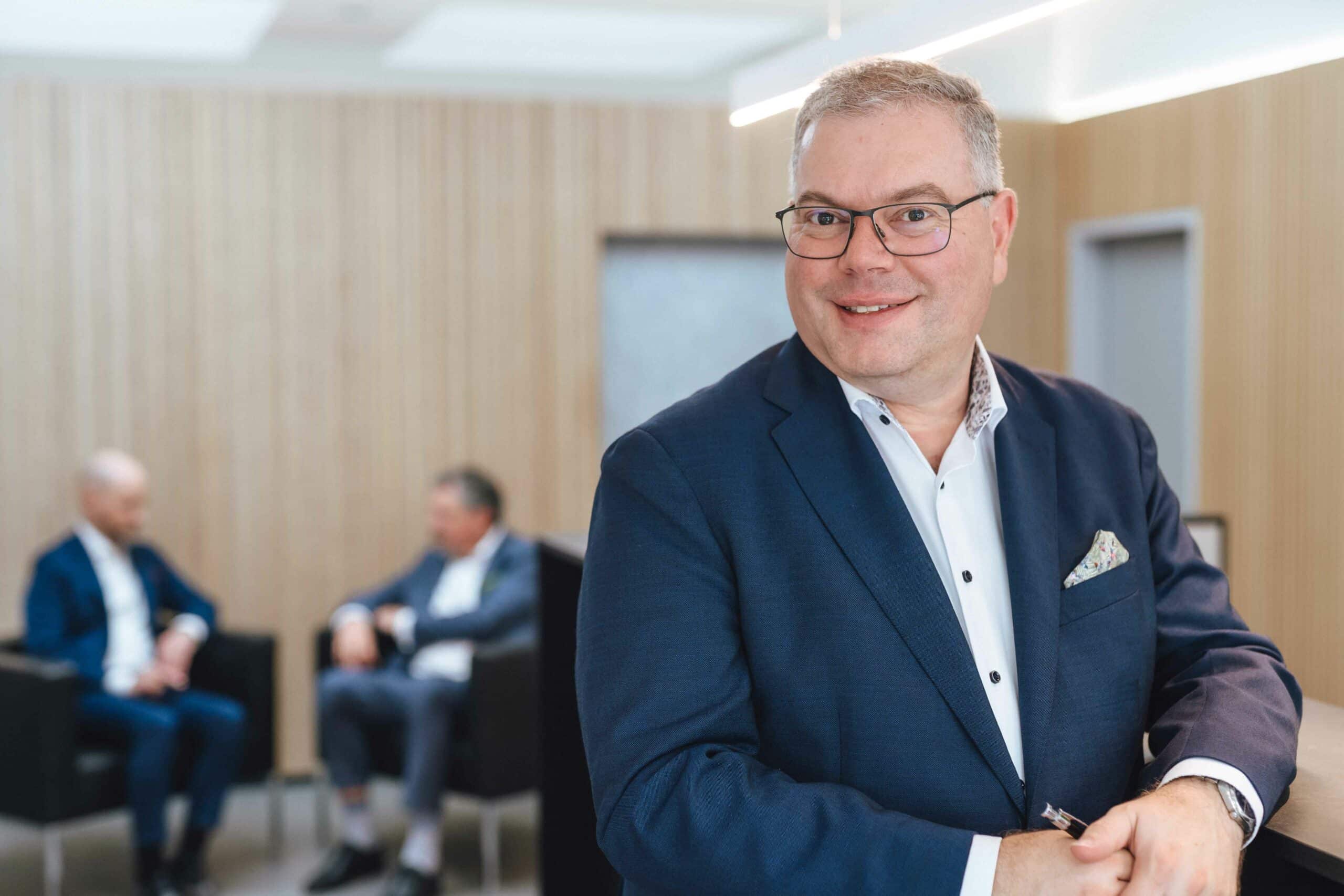 Jan Arnold im Fokus, Kollegen im Hintergrund – Notar Jan Arnold lächelt in die Kamera, während Dr. Carsten Walter und Peter Wandel im Hintergrund ein Gespräch führen.