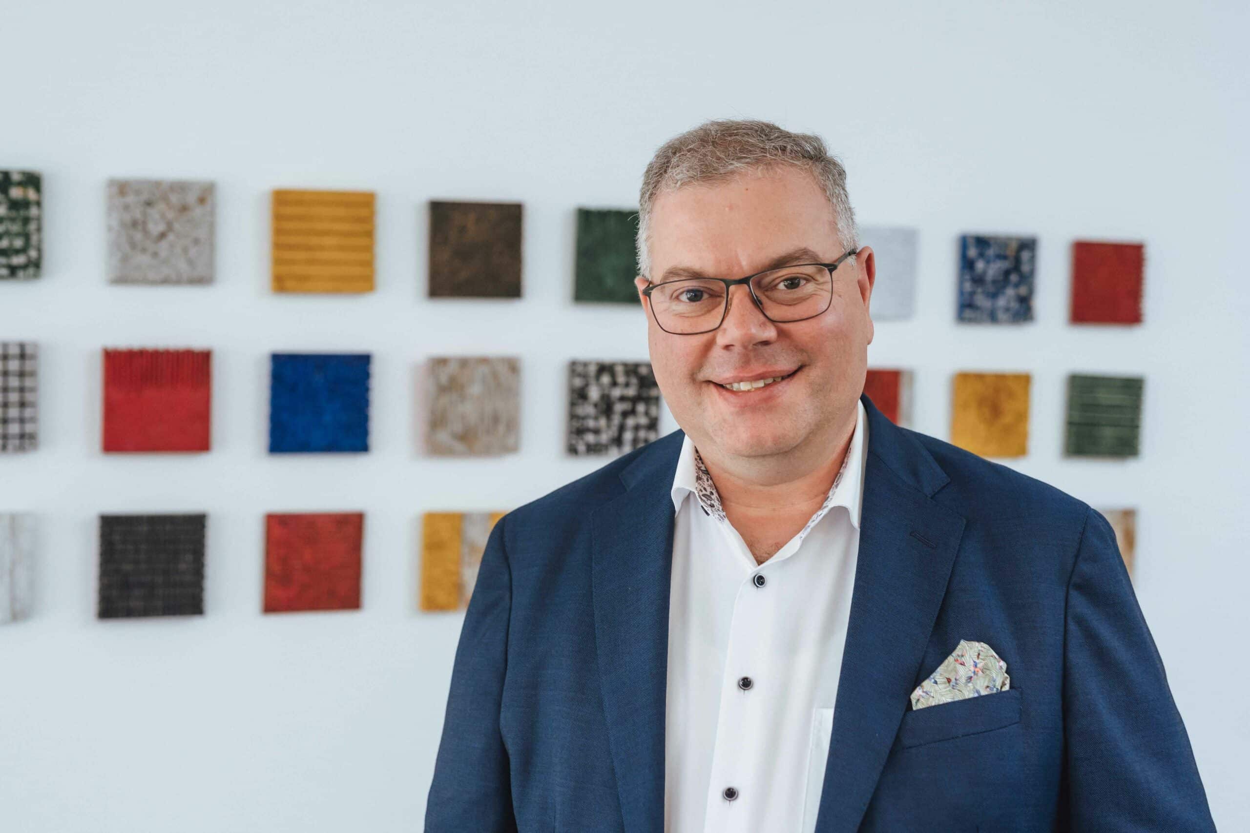 Jan Arnold vor Kunstwand – Notar Jan Arnold steht vor einer weißen Wand mit farbigen Kunstquadraten und blickt lächelnd in die Kamera.